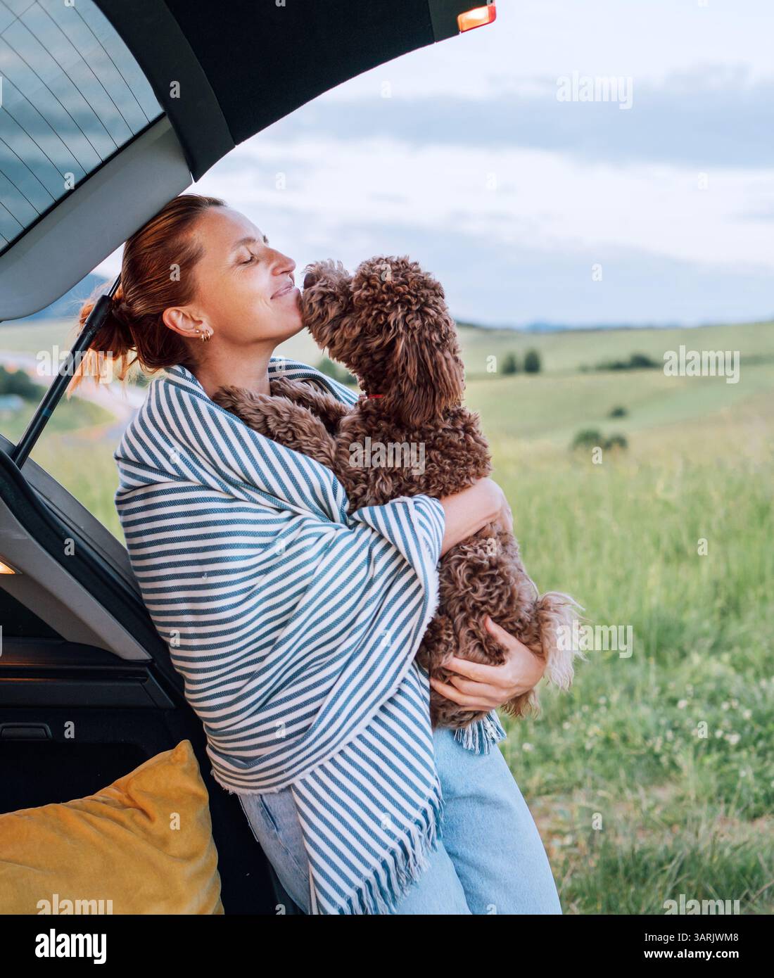 Femme câlin son Maltipoo brun à côté d'un coffre de voiture ouvert, enveloppé dans une couverture rayée, au milieu d'un paysage pittoresque. La scène capture chaud, aff Banque D'Images