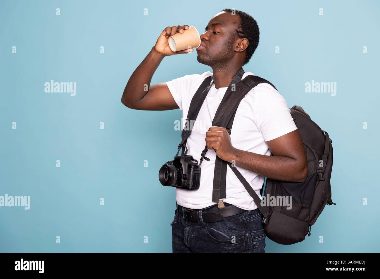Photographe tenant son sac sur son épaule et buvant du café dans un studio. Homme noir portant un t-shirt blanc avec bretelles, un appareil photo autour du cou, saisit son sac à dos et une tasse jetable. Banque D'Images