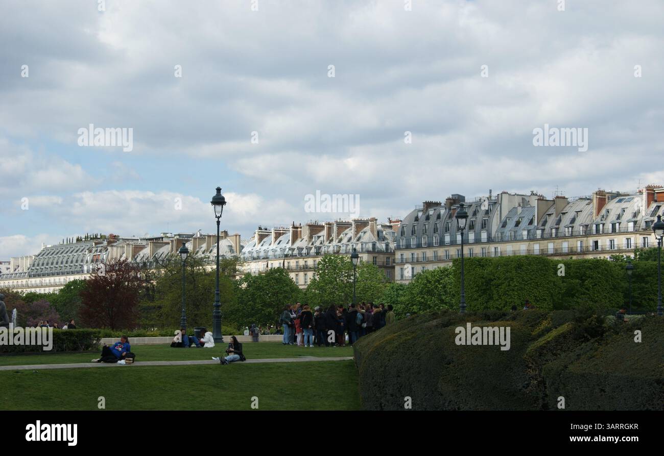 Les gens se détendent sur les pelouses verdoyantes du jardin des Tuileries à Paris, avec une architecture parisienne classique formant une toile de fond pittoresque sous un ciel nuageux. Banque D'Images
