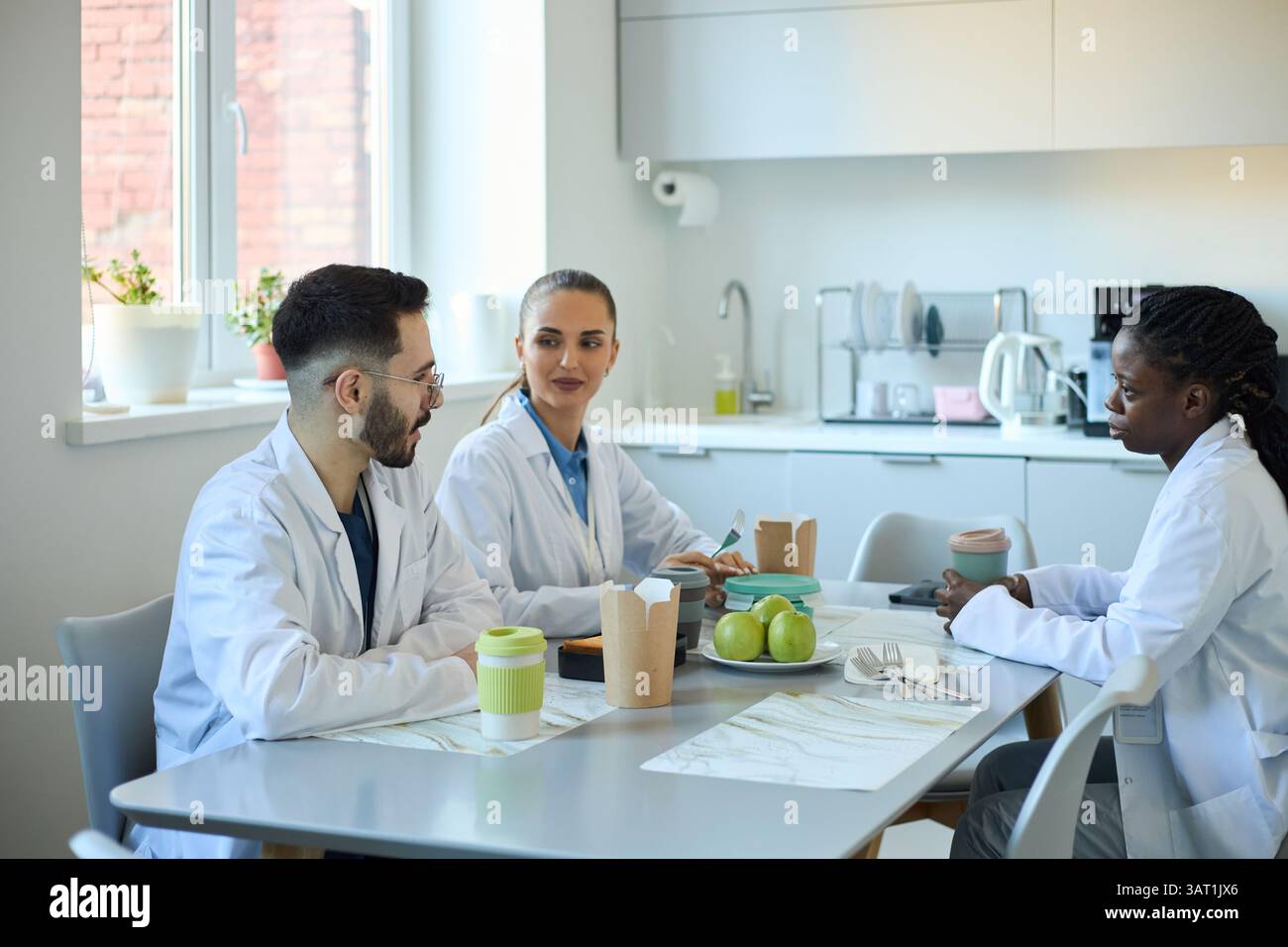 Groupe de trois professionnels de la santé discutant du travail assis autour d'une table dans la salle de pause moderne. Ils partagent des idées autour du café et des collations, fo Banque D'Images