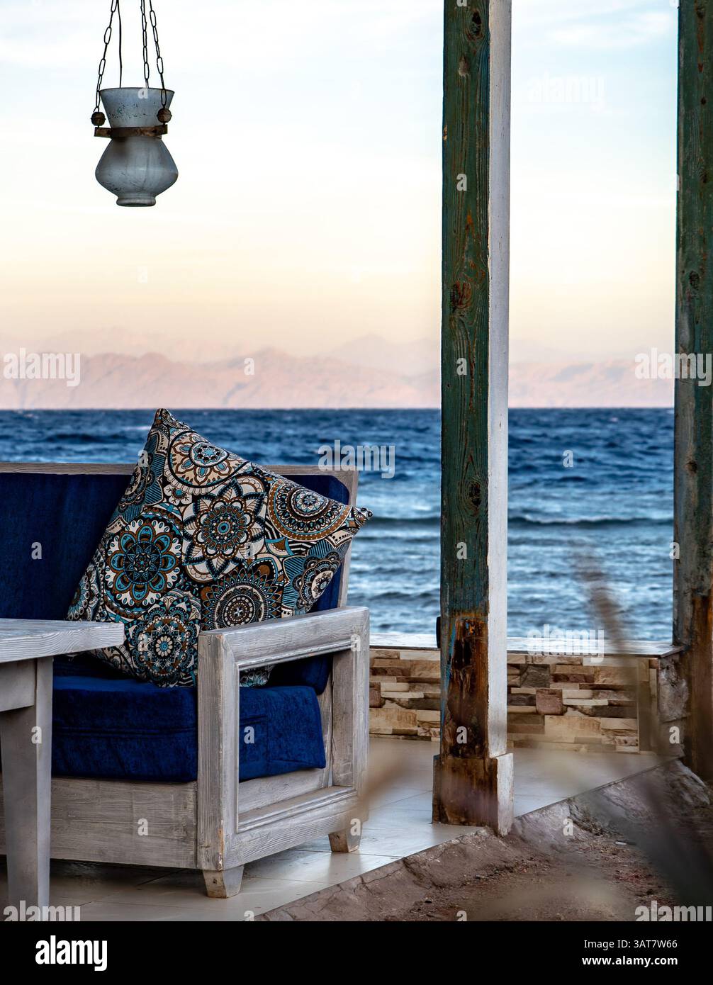 Un coin salon extérieur confortable avec un oreiller décoratif sur une chaise en bois, donnant sur une vue sereine sur l'océan. La scène présente une lanterne suspendue à un p. Banque D'Images