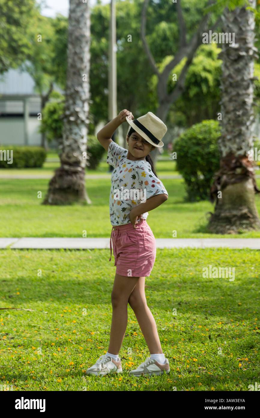 Une jeune fille se tient debout dans un parc, portant un chapeau de paille et un short rose. Elle sourit et pose pour une photo Banque D'Images Une jeune fille se tient debout dans un parc, portant un chapeau de paille et un short rose. Elle sourit et pose pour une photo Banque D'Images
