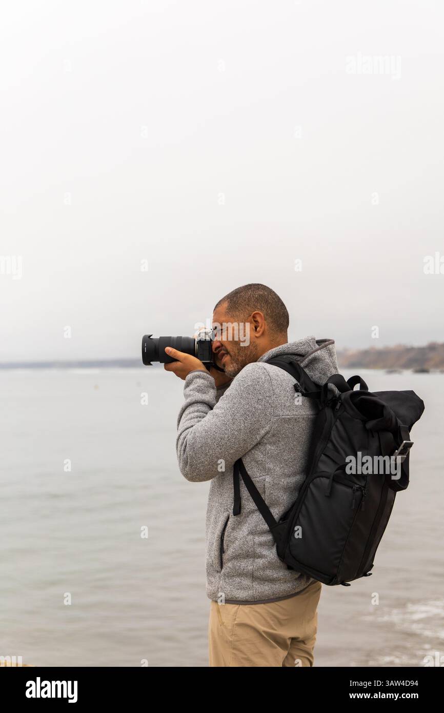 Un homme avec un appareil photo est debout sur une plage, portant un sac à dos. Il prend une photo de l'océan Banque D'Images Un homme avec un appareil photo est debout sur une plage, portant un sac à dos. Il prend une photo de l'océan Banque D'Images