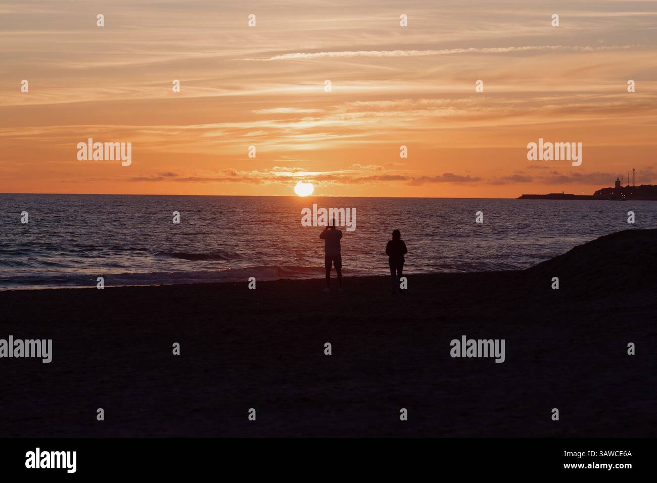 Couple marchant ensemble, silhouetté contre un coucher de soleil orange vibrant, debout pieds nus sur le rivage sablonneux avec des vagues océaniques tranquilles Banque D'Images