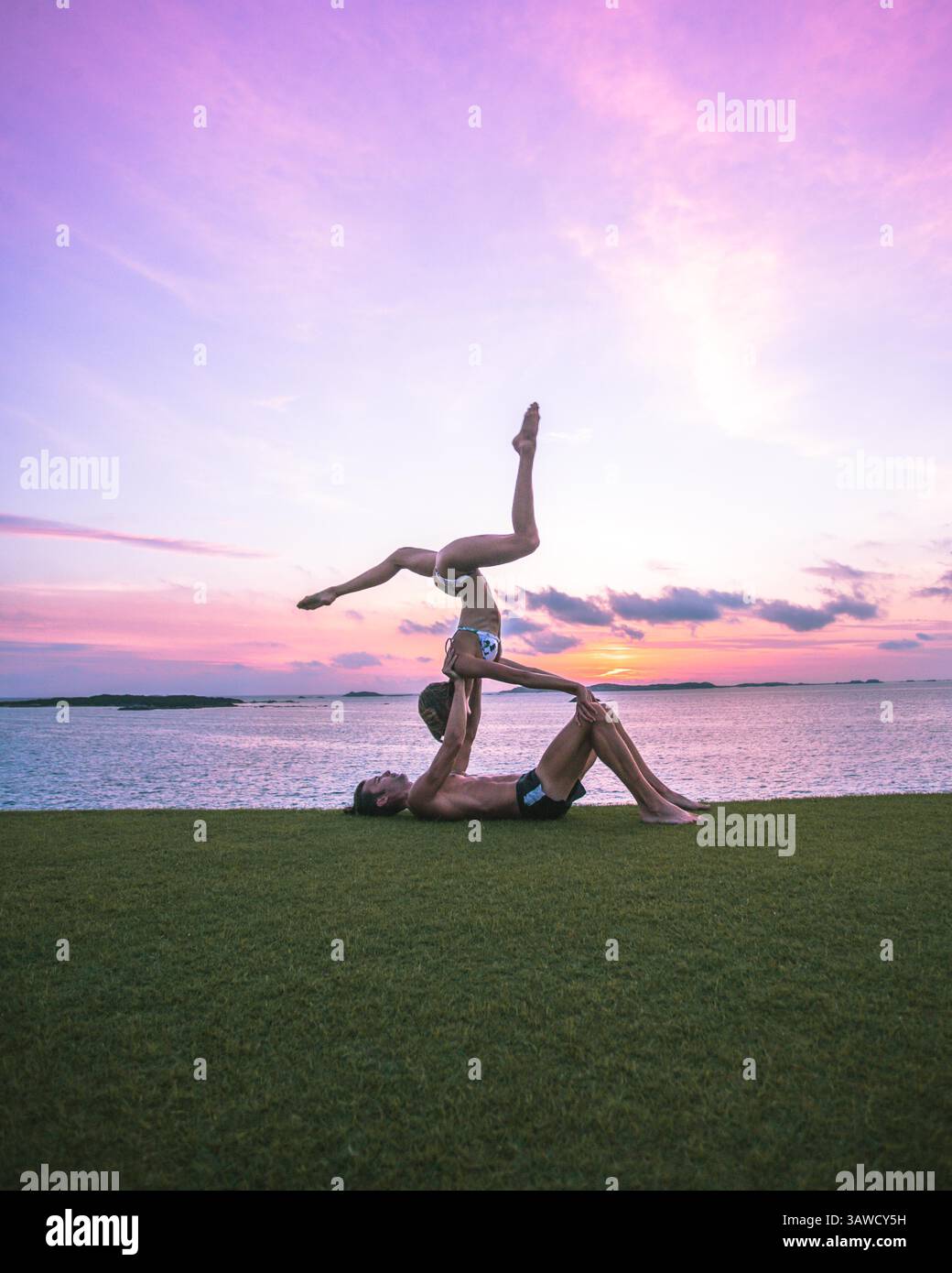 Couple sportif silhouettes homme et femme faisant une pose de yoga acrobatique sur l'herbe devant un beau coucher de soleil sur la mer. Banque D'Images