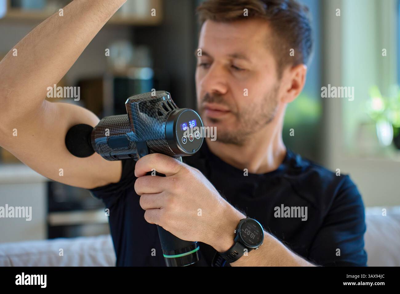 Homme athlétique utilisant un dispositif de massage à percussion à l'intérieur. L'homme applique un pistolet de massage au muscle du bras tout en étant assis dans le salon, thérapie de récupération après l'exercice Banque D'Images