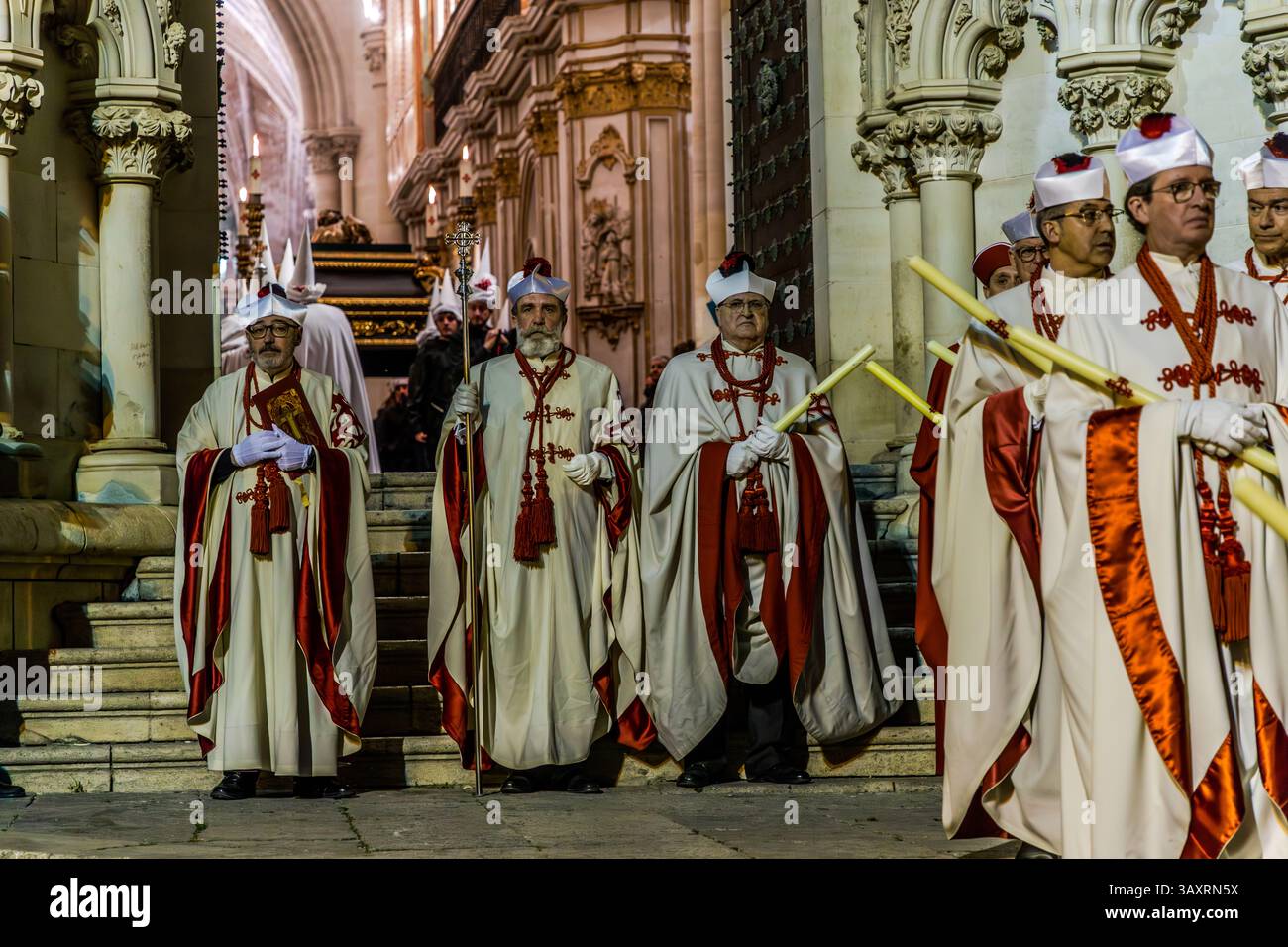 Hommes en vêtements liturgiques blancs-rouges festifs et en hautes mitres, peut-être des chanoines de cathédrale, attendant avec des bâtons de bougies allumés à l'intérieur de l'église à Cuenca, un autre paso visible sur une plateforme étagée en arrière-plan. Calle San Pedro, Cuenca, Castilla-La Mancha, Espagne. Pendant la Semana Santa à Cuenca, 33 Hermandades participent à onze processions à travers la ville pendant la Semaine Sainte. Banque D'Images