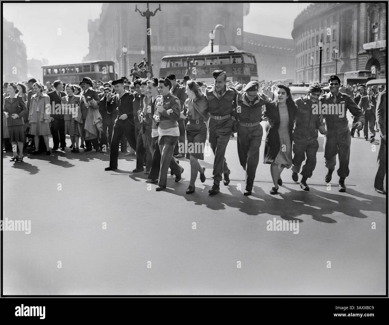 VJ DAY LONDON les civils britanniques et le personnel de service du Piccadilly Circus de Londres célèbrent la nouvelle de la victoire alliée sur le Japon en août 1945. LA FIN DE LA seconde Guerre mondiale dans Piccadilly Circus à Londres, un groupe d'hommes et de femmes militaires, se lient les bras alors qu'ils marchent vers la caméra, chantant et dansant dans la célébration autour d'Eros, sur la nouvelle que la guerre au Japon est terminée. Derrière eux, des foules de gens se rassemblent au soleil. Plusieurs bus peuvent également être vus. Vue de la statue d'Eros, vers Piccadilly & Regent Street. Londres Royaume-Uni date 1945 Banque D'Images