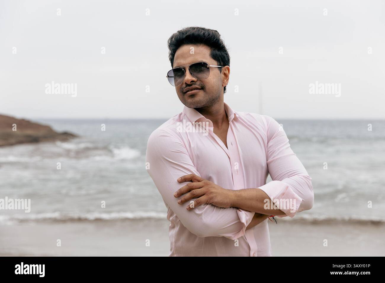 Homme confiant en lunettes de soleil avec bras pliés debout par l'océan. Banque D'Images