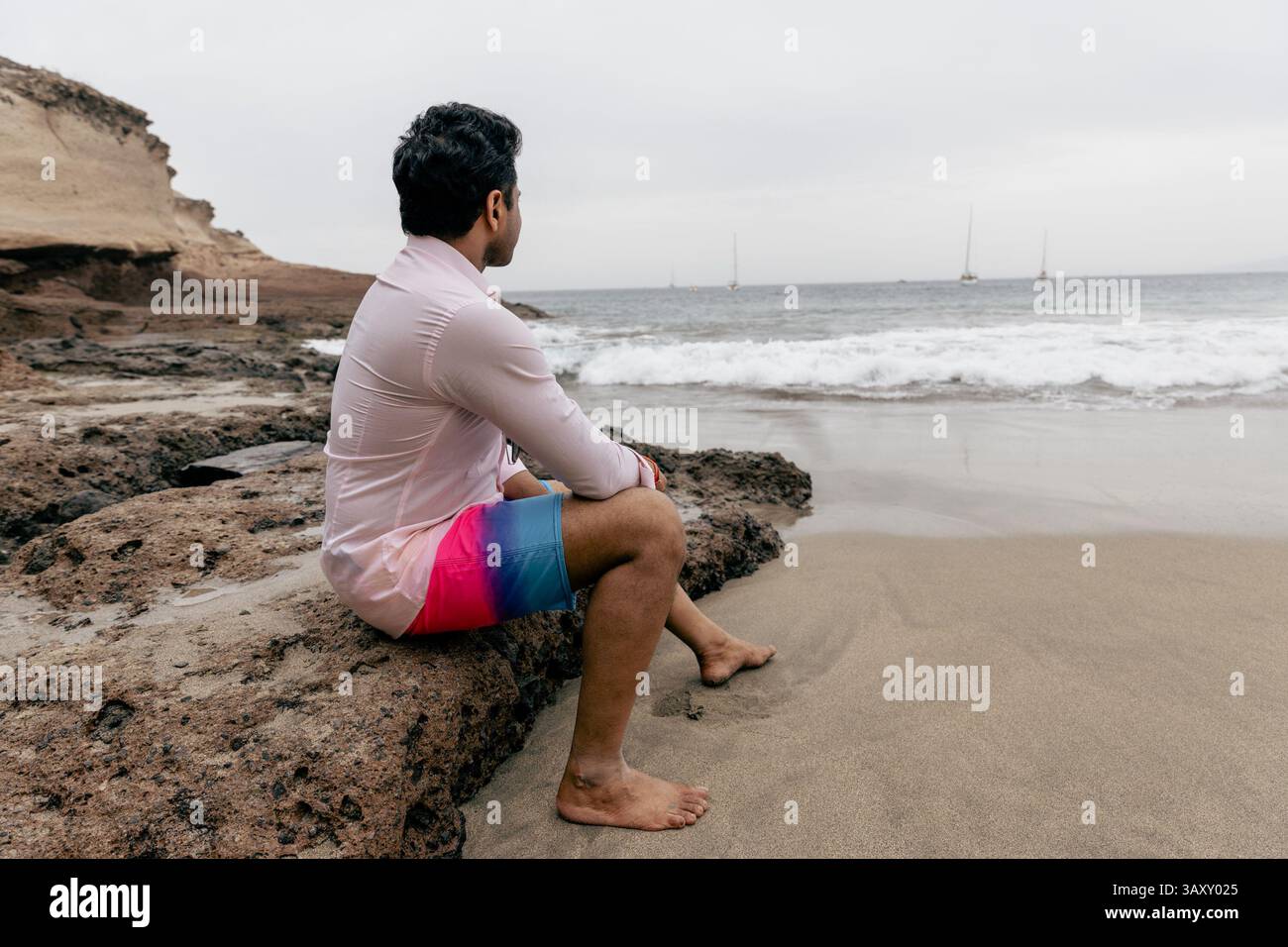 Un homme assis sur un rocher à la plage regardant l'océan et se relaxant. Banque D'Images