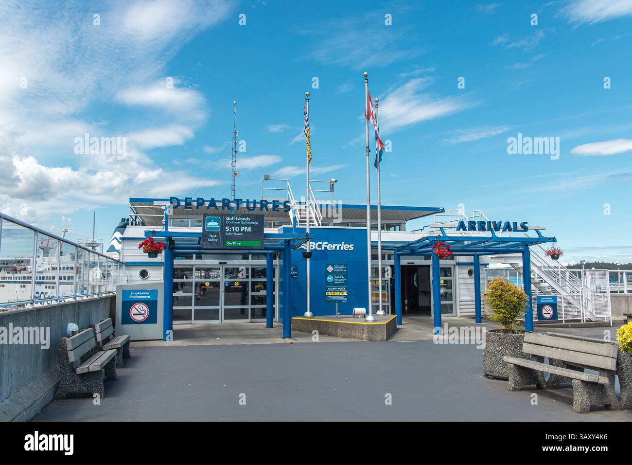 BC Ferries Swartz Bay terminal entrée montrant des panneaux d'arrivée et de départ sous un ciel lumineux, avec des drapeaux et des panneaux visibles pour les voyageurs. Banque D'Images