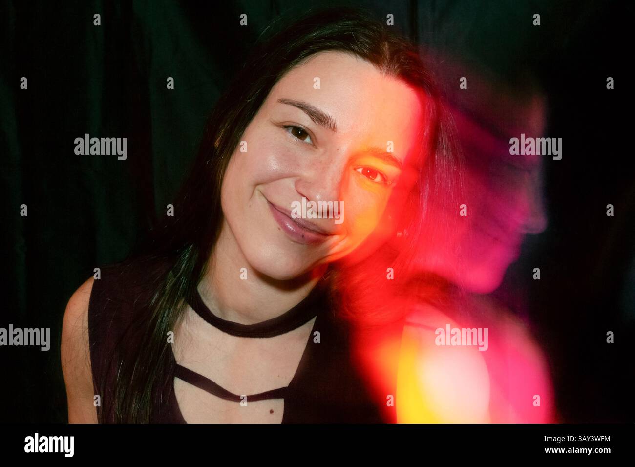 Une femme sourit chaleureusement, son visage mis en évidence par des effets de lumière rouge vif sur un fond sombre, créant un portrait moderne et artistique avec un vif Banque D'Images