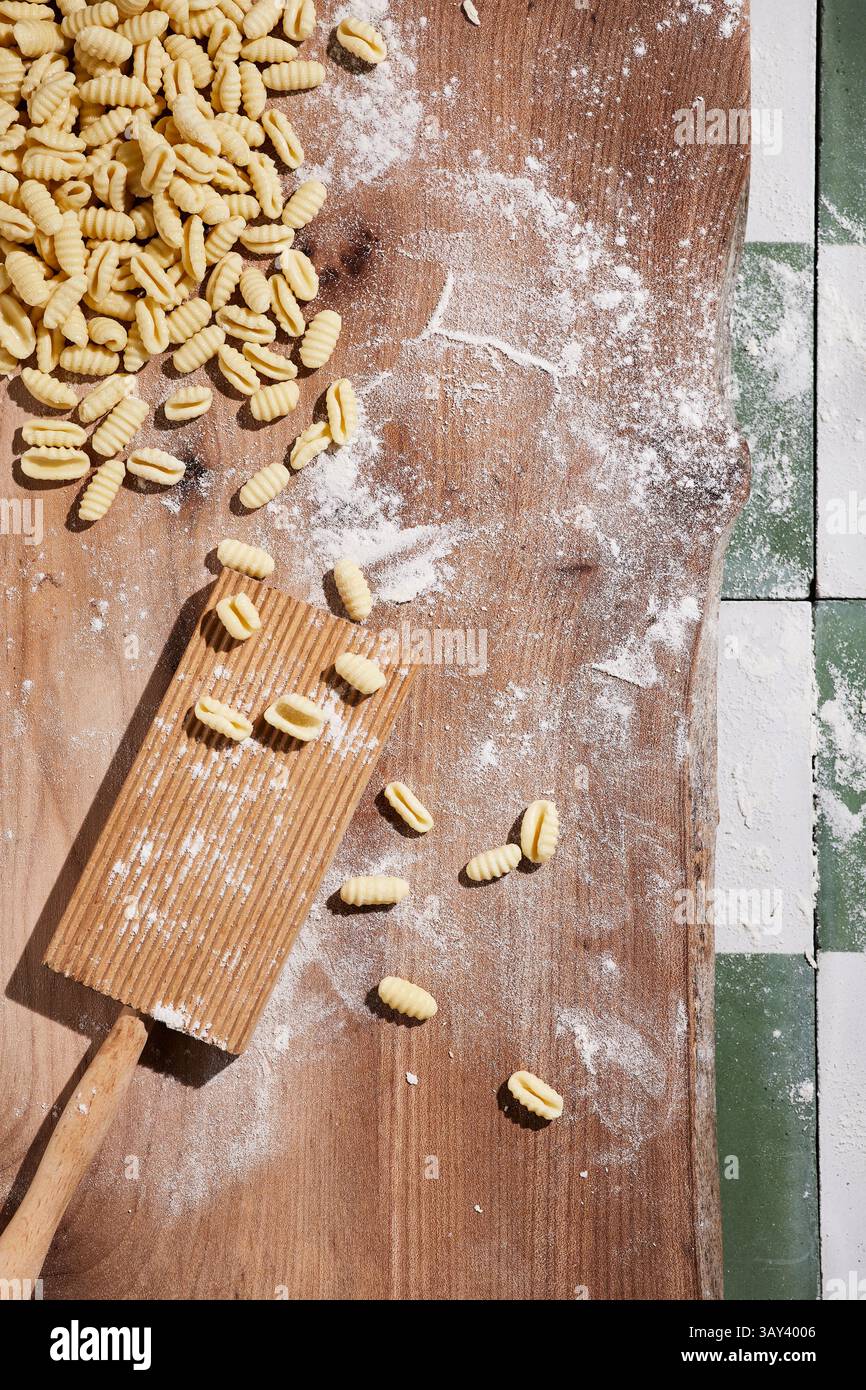 Vue de dessus de gnocchi maison frais dispersés sur une surface en bois saupoudrée de farine à côté d'une planche à gnocchi et d'un rouleau à pâtisserie, soulignant le processus du passé Banque D'Images