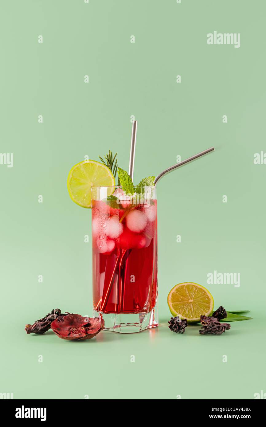 Un verre vibrant de thé glacé hibiscus orné de tranches de citron vert, de feuilles de menthe et de glaçons, sur fond vert pastel, offrant un rafraîchissement Banque D'Images