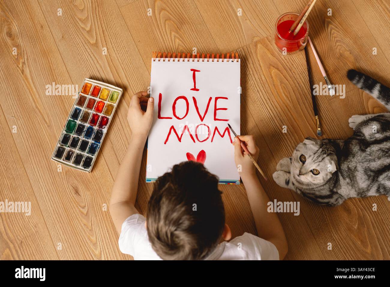 Un enfant peint « I LOVE MAMAN » sur un carnet à l’aquarelle rouge, tandis qu’un chat curieux est allongé à côté d’eux. Entouré de pinceaux et de peintures, la scène cap Banque D'Images