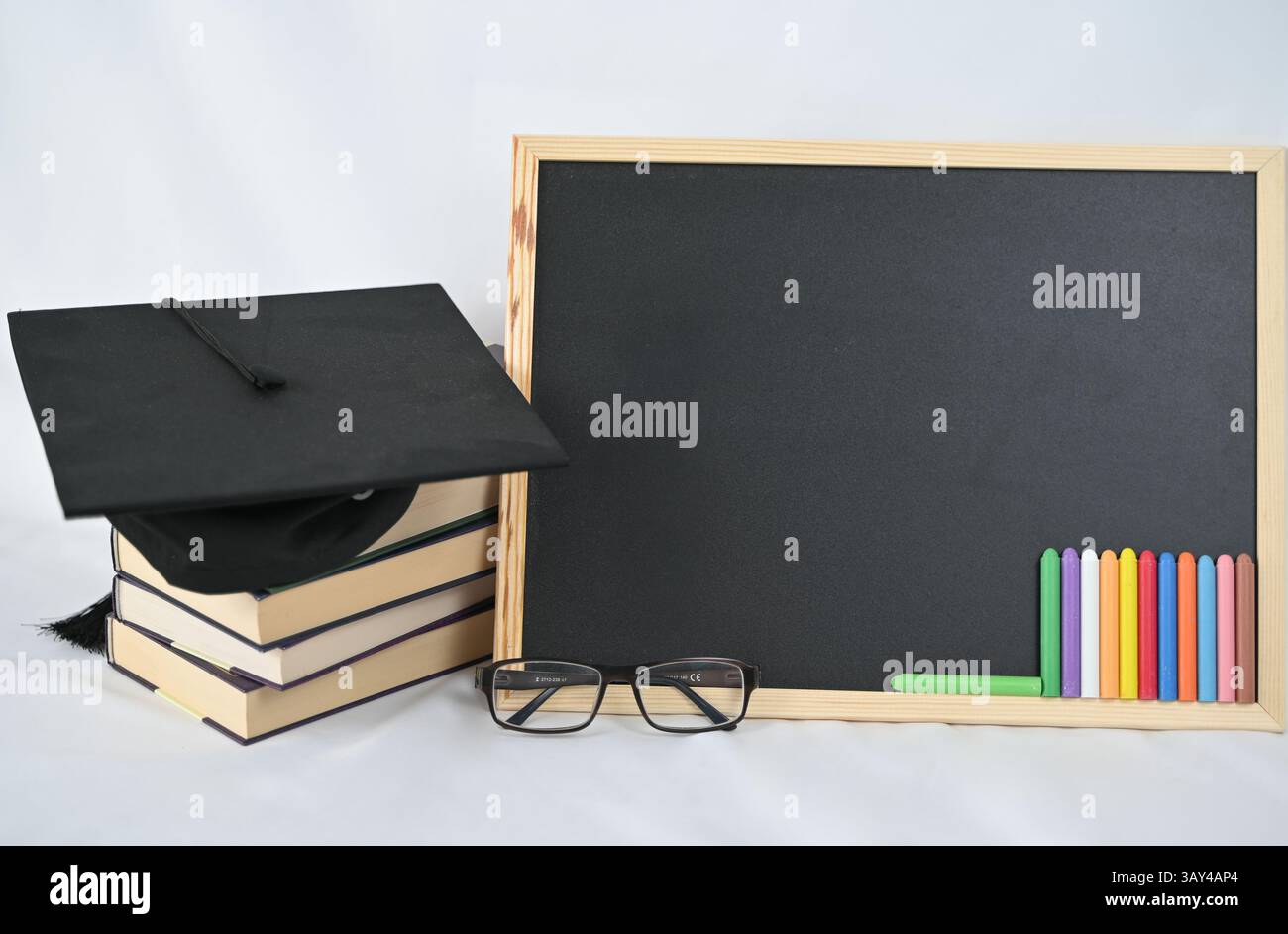 Une casquette de graduation est perchée sur une pile de livres, positionnée juste à côté d'un tableau noir et d'une paire de lunettes pour la lecture Banque D'Images