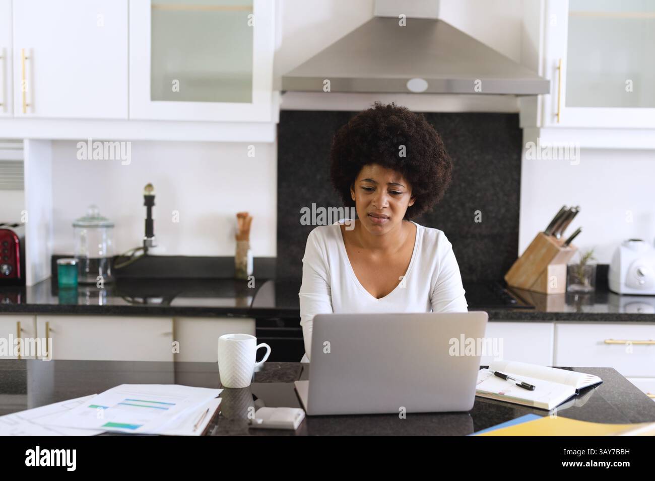 Femme afro-américaine travaillant sur ordinateur portable à l'îlot de cuisine moderne, avec des documents et une tasse à café Banque D'Images