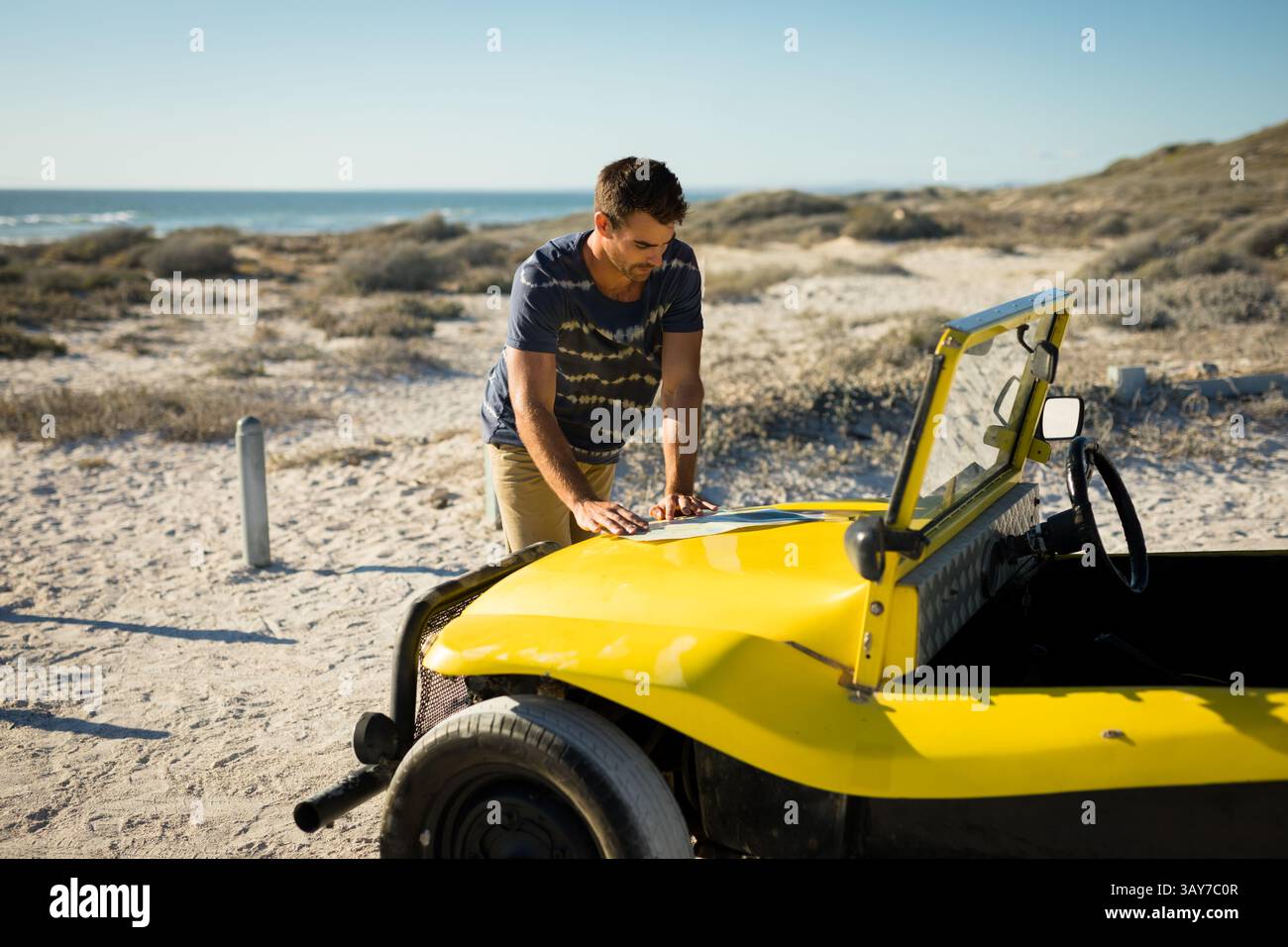 Homme penchant et inspectant le capot du buggy dune jaune sur la dune côtière, avec vue sur l'océan Banque D'Images