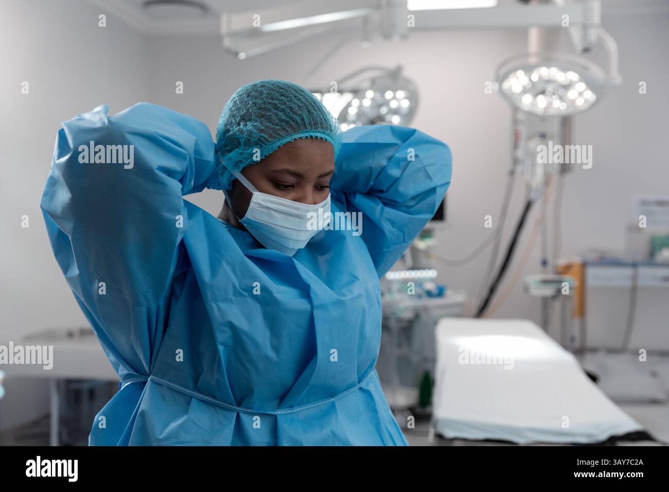 Jeune femme afro-américaine ajustant la robe avec bonnet de bouffant et masque dans la salle d'opération Banque D'Images