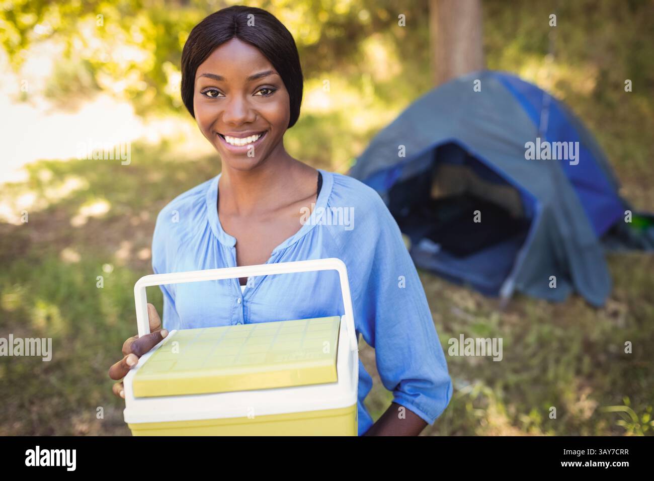 Jeune femme afro-américaine tenant une glacière près de la tente de camping au camping, profitant d'une journée ensoleillée Banque D'Images