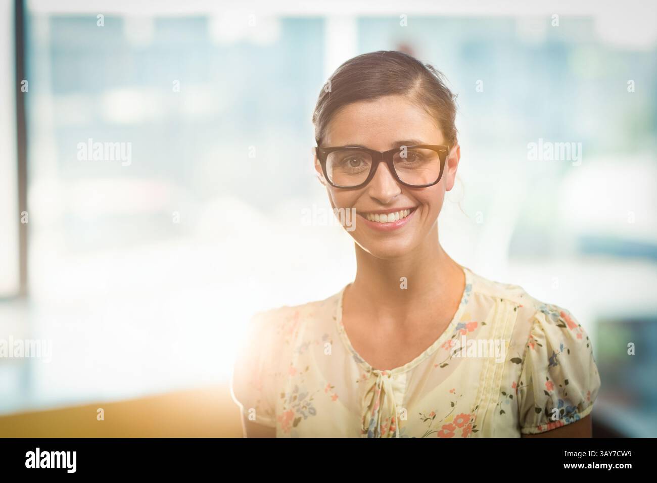 Jeune femme adulte debout et souriante au bureau avec des lunettes encadrées en noir et un chemisier floral Banque D'Images