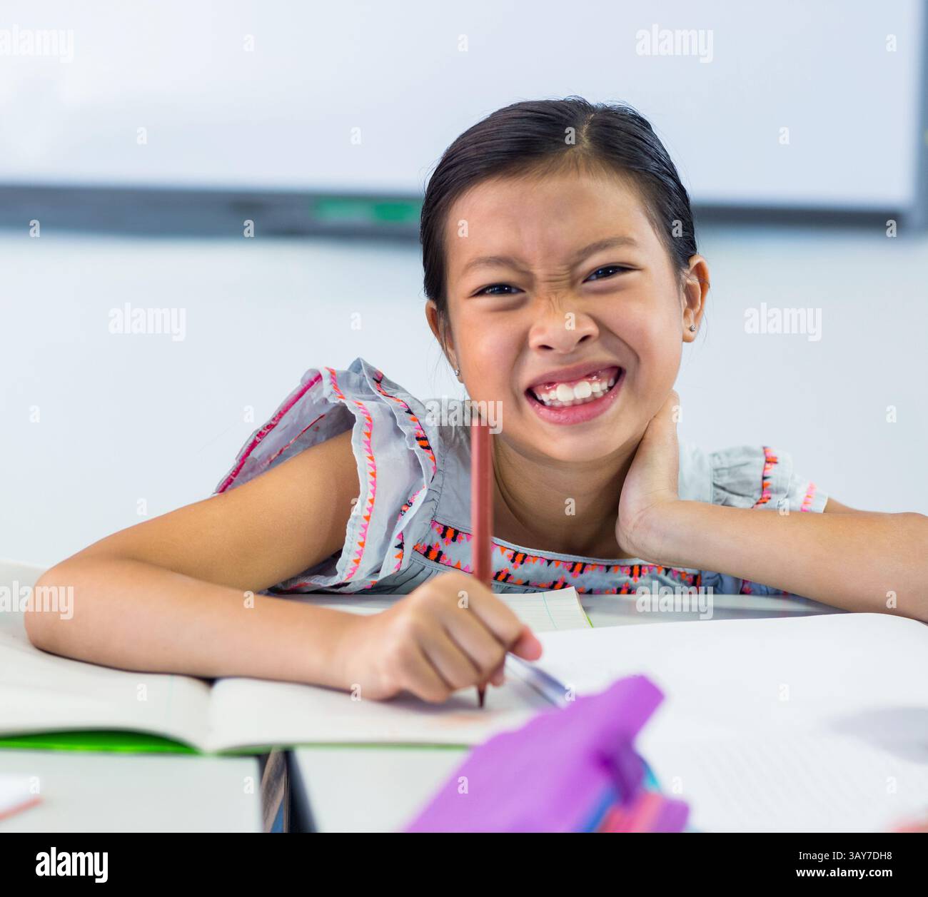 Fille asiatique écrivant au bureau de classe de l'école avec des cahiers ouverts, crayon et tableau blanc derrière Banque D'Images