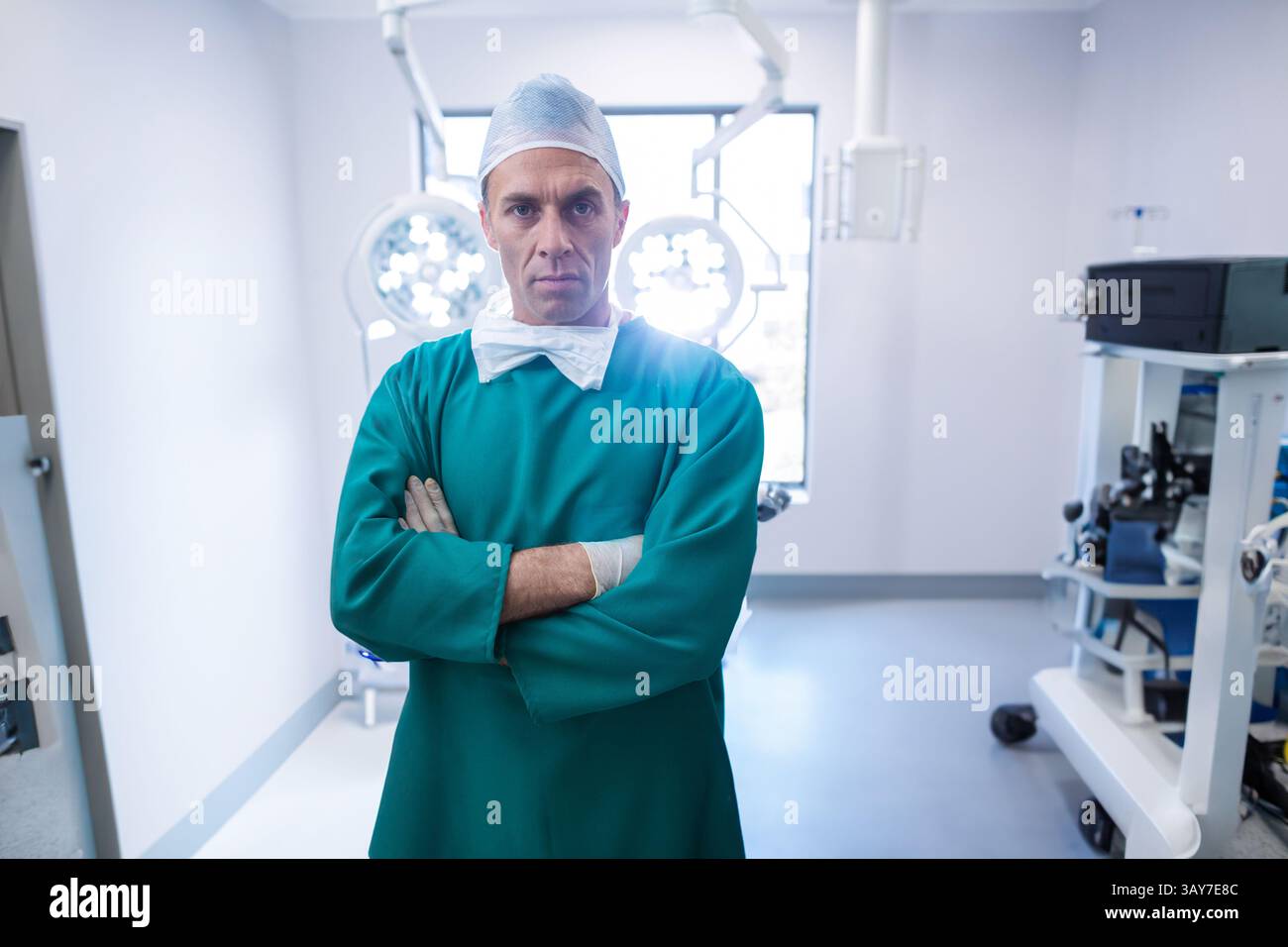 Chirurgien masculin debout, les bras croisés dans la salle d'opération, avec des lampes chirurgicales et un chariot d'équipement Banque D'Images