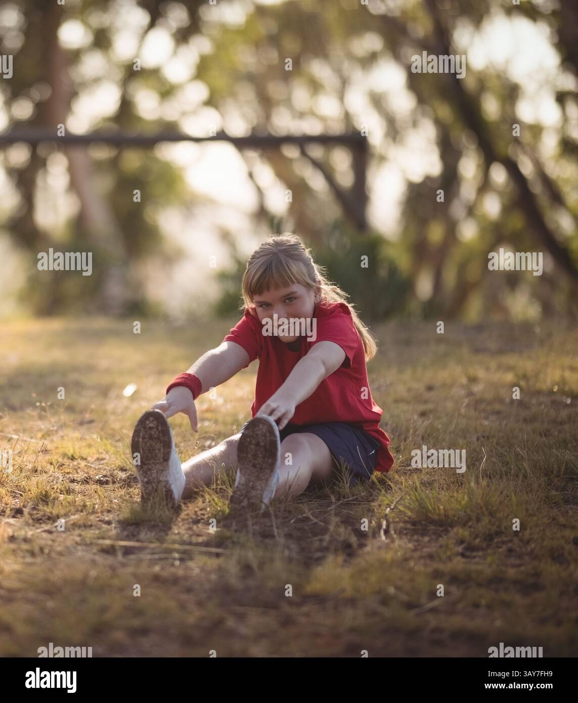 Enfant s'étirant pour atteindre les orteils dans les bois ensoleillés, avec bracelet rouge et baskets blanches Banque D'Images Enfant s'étirant pour atteindre les orteils dans les bois ensoleillés, avec bracelet rouge et baskets blanches Banque D'Images