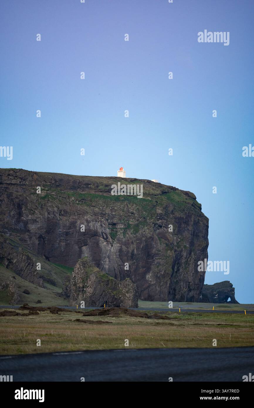 Phare au sommet d'une falaise spectaculaire près de Dyrhólaey, Islande, sous un ciel bleu clair au crépuscule, avec un premier plan herbeux et des formations rocheuses volcaniques. Banque D'Images