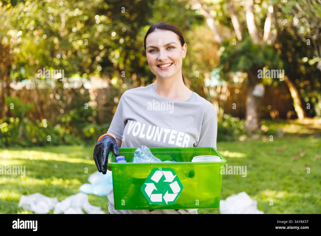 Femme bénévole tenant un bac de recyclage vert dans le parc ensoleillé, souriant avec des bouteilles et des sacs en plastique Banque D'Images