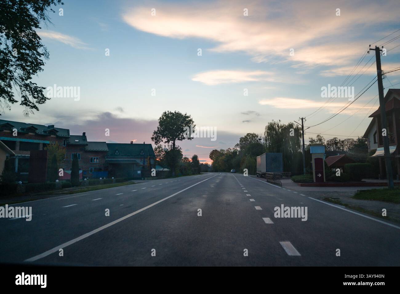 Alors que le crépuscule s'installe, une route rurale tranquille s'étend devant vous, encadrée par de charmantes maisons. Les douces teintes pastel dans le ciel donnent une ambiance paisible à la sérénité Banque D'Images