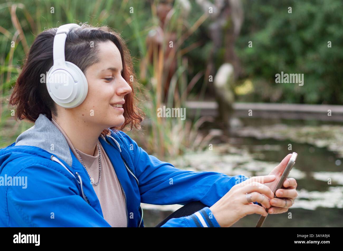 Heureuse jeune femme portant des écouteurs sans fil choisit la musique de playlist sur son application smartphone, assise dans un parc avec étang et arbres à l'arrière Banque D'Images