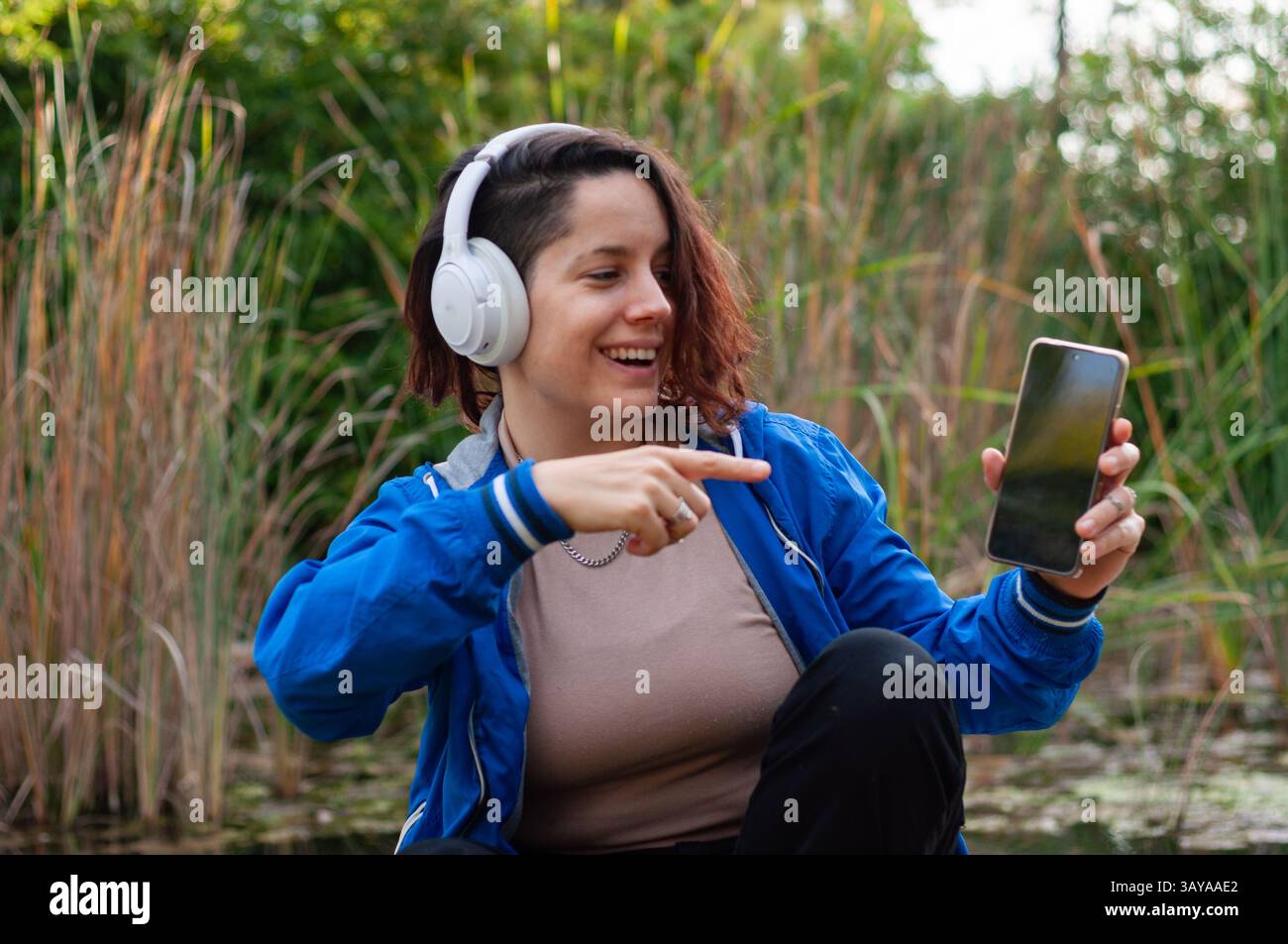 Assise près d'un étang, une jeune femme portant des écouteurs profite d'une application de streaming de musique sur son téléphone, pointant vers l'écran et souriant Banque D'Images
