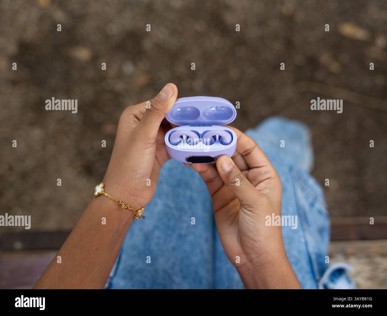 Femme assise à l'extérieur, tenant ouvert un étui de charge d'écouteurs sans fil violet, révélant les écouteurs à l'intérieur, profitant d'un moment de loisirs et de relaxatio Banque D'Images