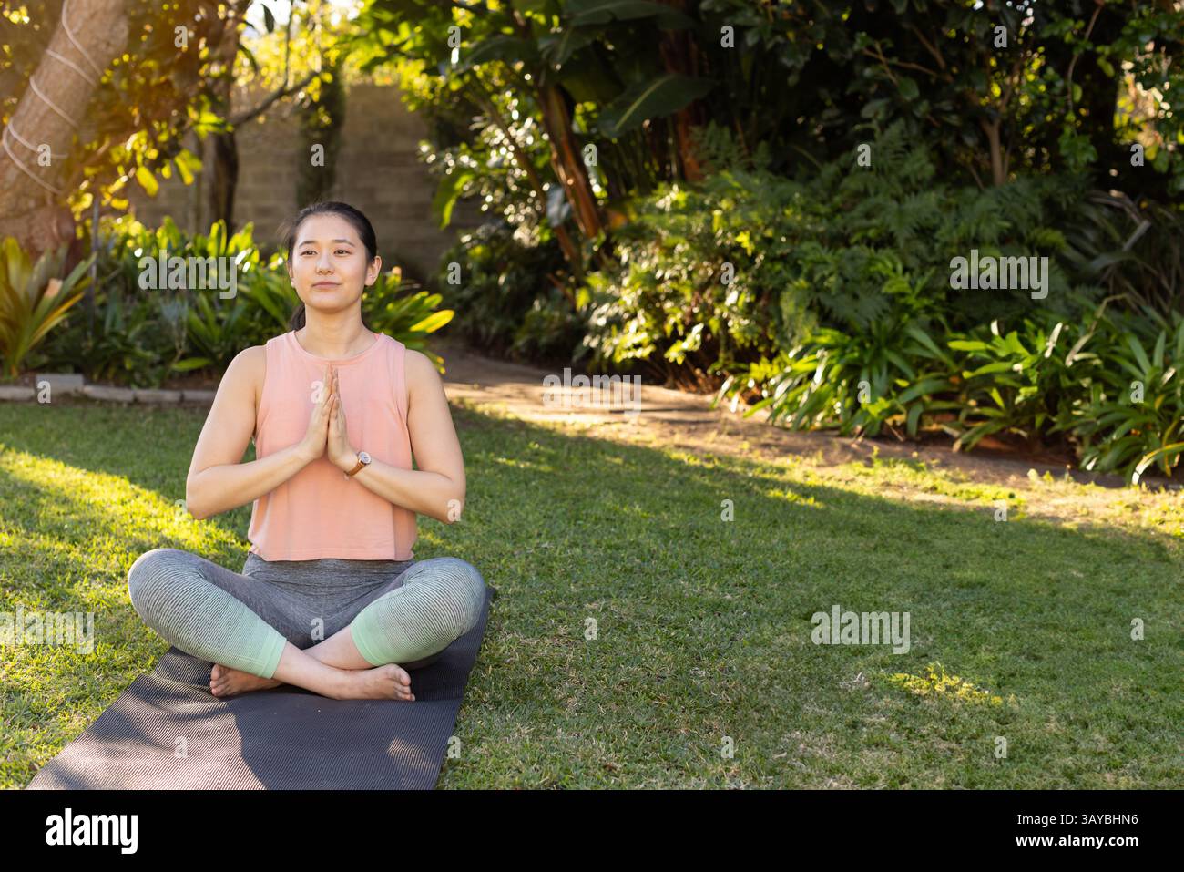 Femme asiatique pratiquant la croix de yoga ?legged sur tapis de yoga dans le jardin de la maison, mettant en valeur la lumière du soleil tapissée Banque D'Images