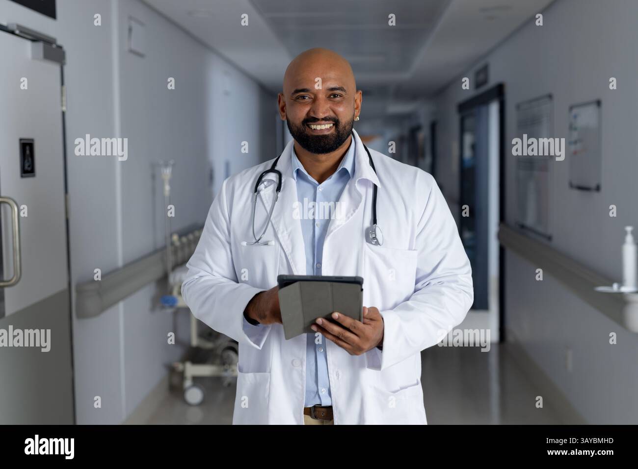 Médecin masculin tenant la tablette et souriant dans le couloir de l'hôpital, avec stéthoscope et statif IV Banque D'Images
