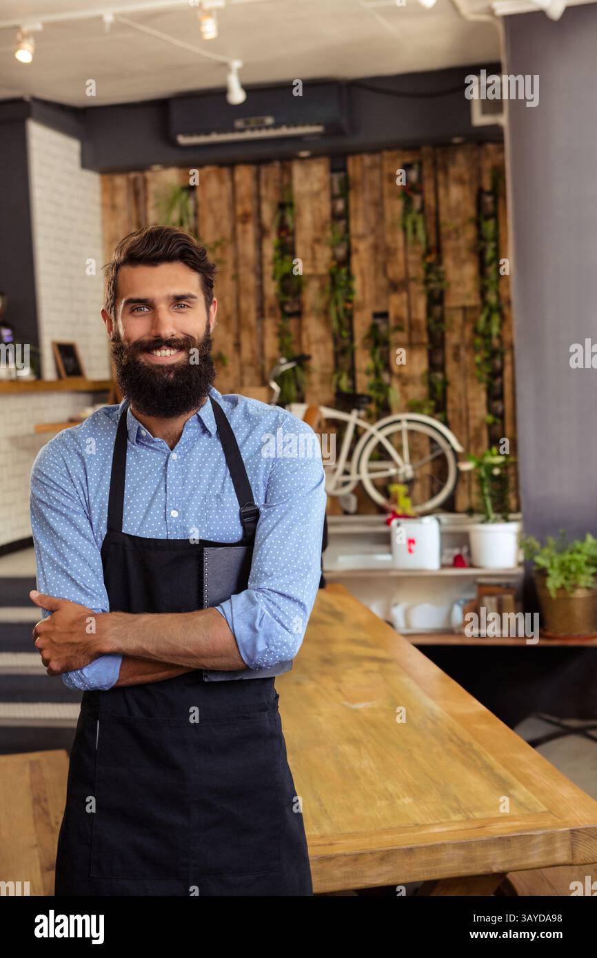 Jeune homme adulte portant un tablier, travaillant derrière un comptoir en bois dans un café confortable, avec art mural vélo Banque D'Images