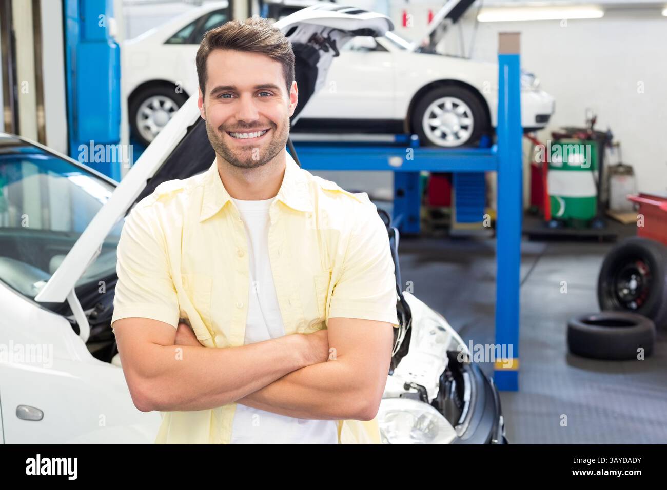 Jeune homme debout bras croisés dans le garage automobile, montrant le capot ouvert de voiture blanche et l'ascenseur hydraulique Banque D'Images