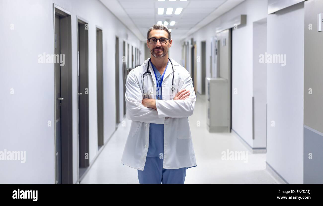 Médecin masculin debout dans le couloir de l'hôpital, portant une blouse de laboratoire et des gommages bleus et tenant le stéthoscope Banque D'Images
