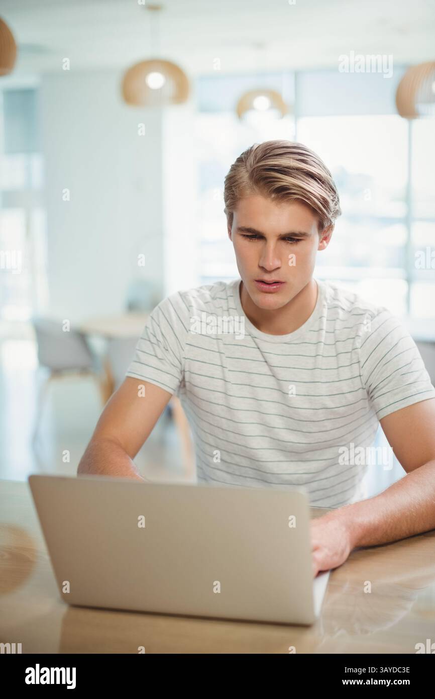 Homme dans la vingtaine travaillant à table dans un espace de travail moderne, avec ordinateur portable argenté et lampes suspendues Banque D'Images