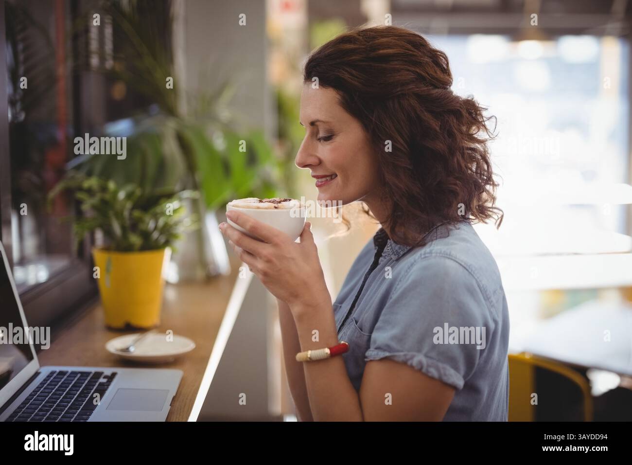 Femme adulte moyenne assise au comptoir de café par la fenêtre tenant le café latte art près du pot jaune Banque D'Images