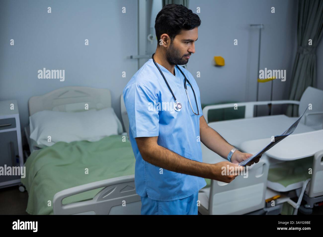 Homme asiatique debout dans la chambre de patient d'hôpital avec des lits, examinant le dossier médical avec stéthoscope Banque D'Images