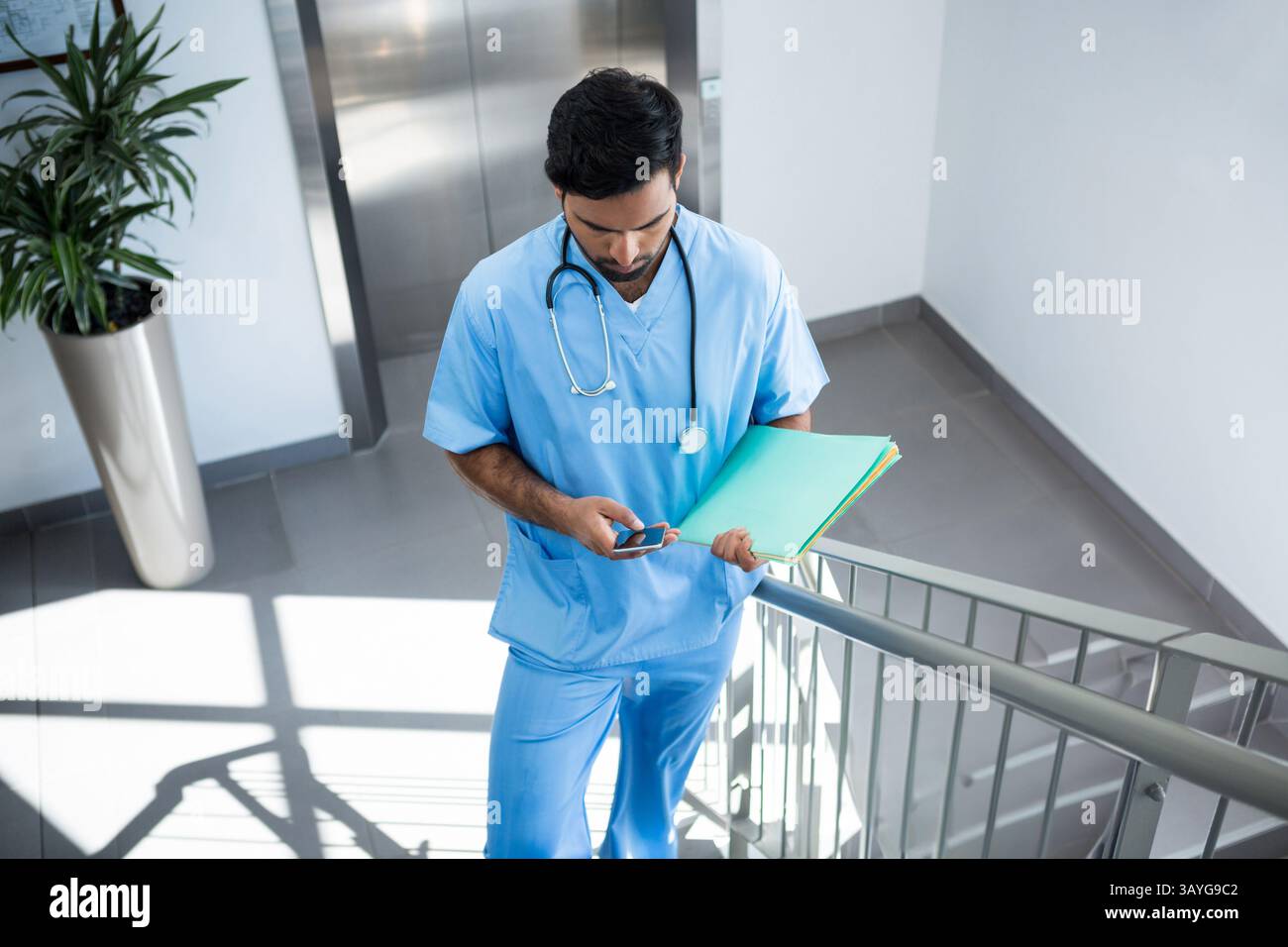 Homme asiatique descendant cage d'escalier de l'hôpital à l'aide d'un smartphone et portant des dossiers de fichiers avec stéthoscope Banque D'Images
