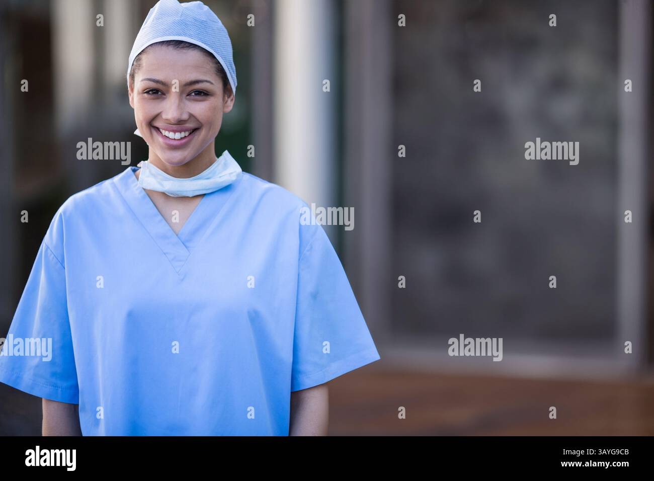 Infirmière debout près de l'entrée de l'hôpital portant des gommages bleu clair et une casquette avec masque autour du cou Banque D'Images