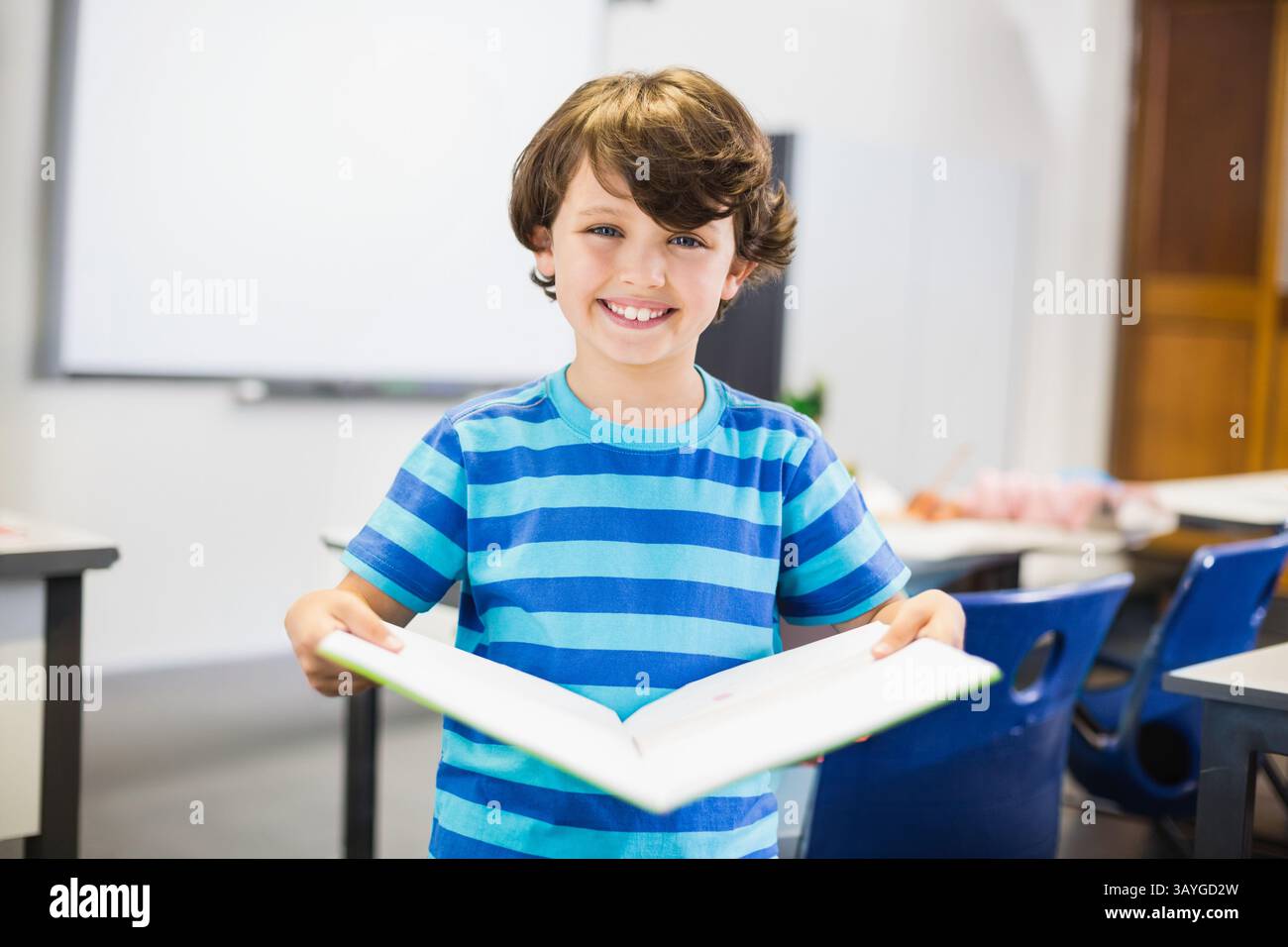 Enfant tenant un livre ouvert à couverture rigide dans la salle de classe de l'école, avec des chaises et des bureaux en plastique bleu Banque D'Images