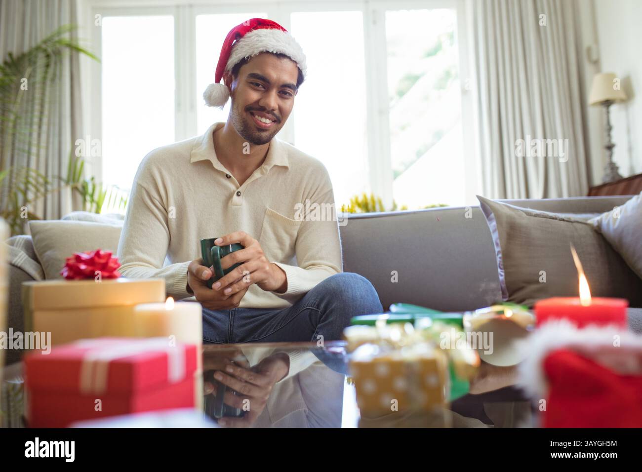 Homme portant chapeau de Père Noël buvant de tasse verte dans le salon, avec des cadeaux emballés et des bougies Banque D'Images
