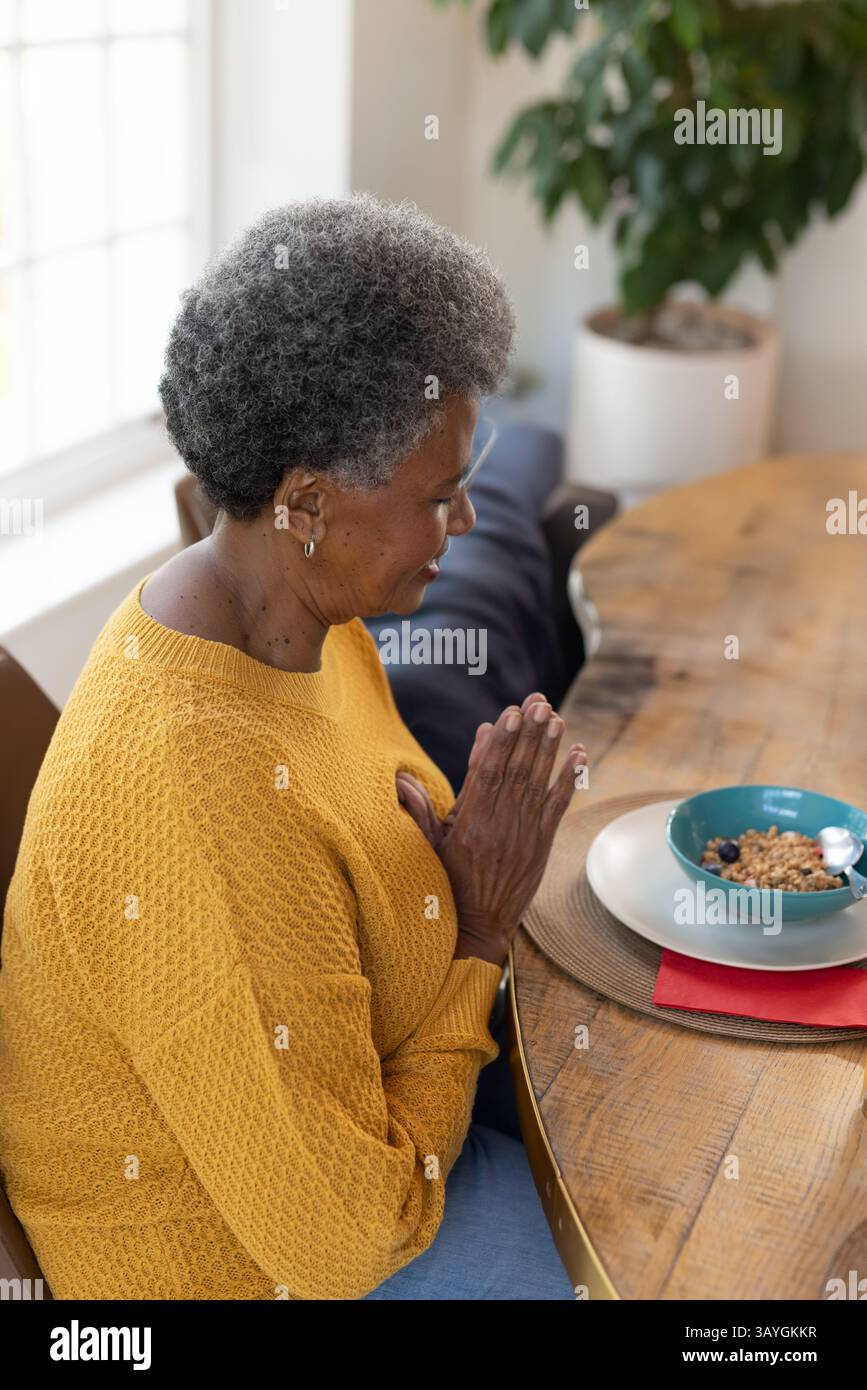 Femme afro-américaine senior priant à la table à manger à la maison, avec des céréales de myrtille, cuillère, plante Banque D'Images