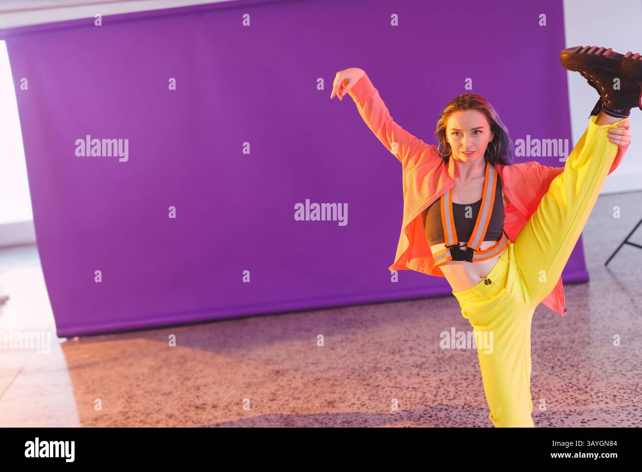 Personne non binaire effectuant une extension de jambe haute en studio, avec support de toile de fond violet Banque D'Images