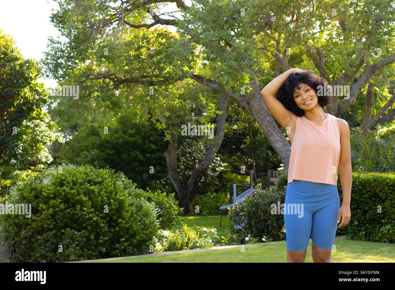 Femme debout sur la pelouse bien entretenue dans le jardin ensoleillé, avec des arbustes verts et banc en bois, espace de copie Banque D'Images