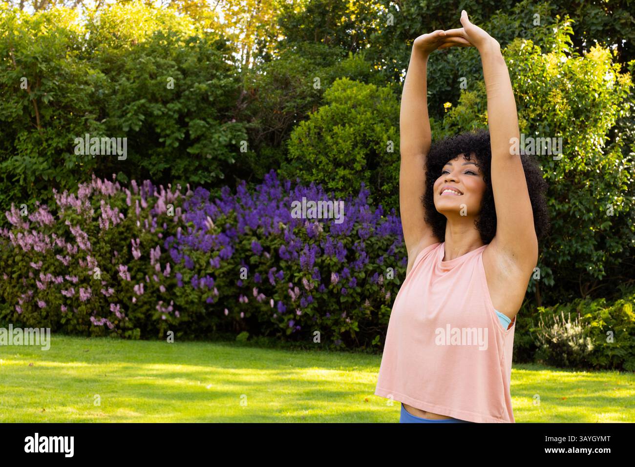Femme étirant les bras au-dessus de la tête sur la pelouse de jardin ensoleillée parmi les arbustes fleuris avec le sourire, espace de copie Banque D'Images